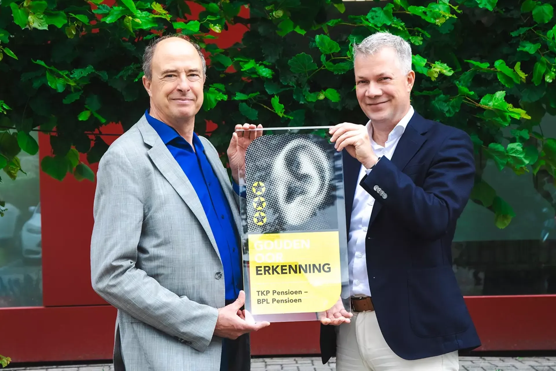 Twee mannelijke bestuurders houden de Gouden Oor Erkenning omhoog voor een groene achtergrond.